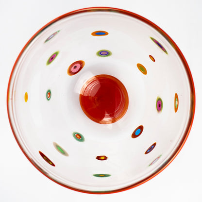 A top-down view of a clear glass bowl with a vibrant orange-red rim, a matching orange-red center, and multiple small multicolored concentric dot patterns scattered across the interior, set against a plain white background.
