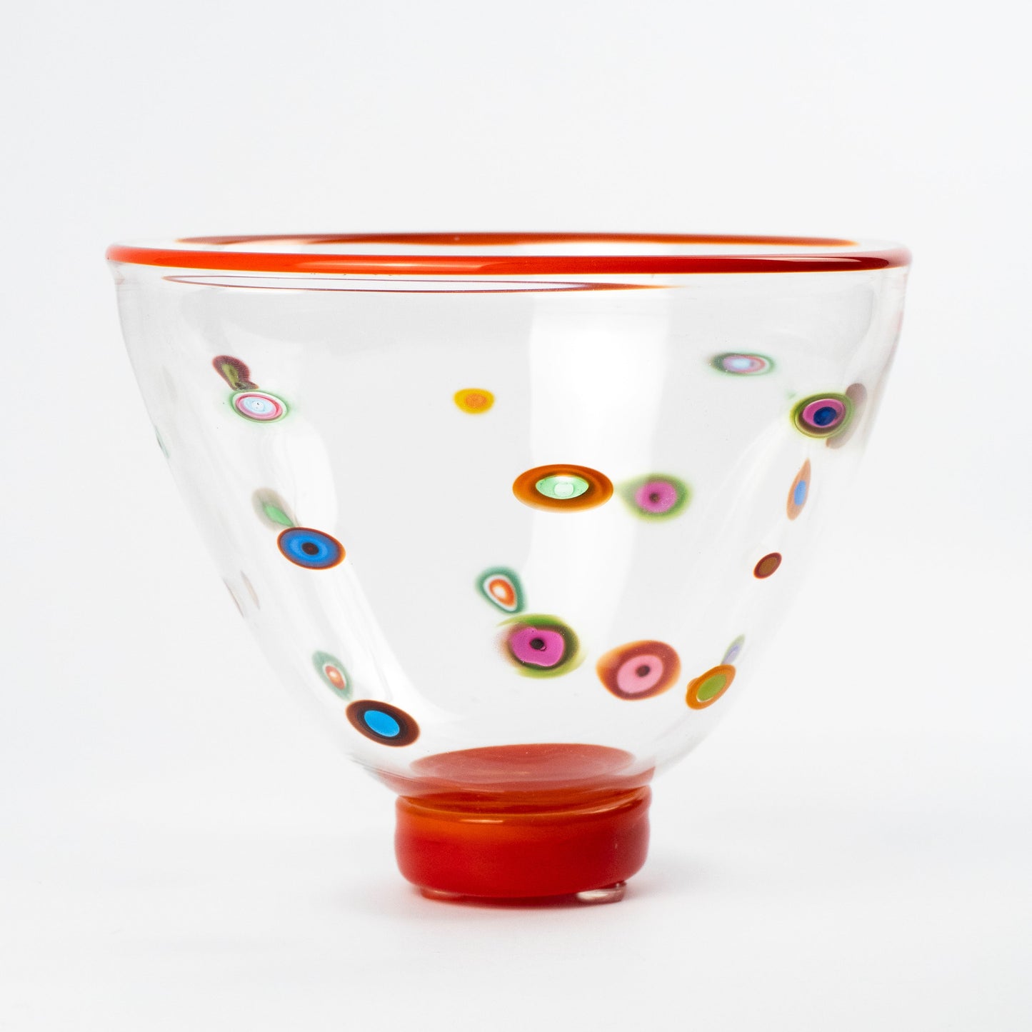 A clear glass bowl with a vibrant red rim and base, decorated with colorful hand-blown glass dots known as Murrine in various patterns, set against a plain white background.