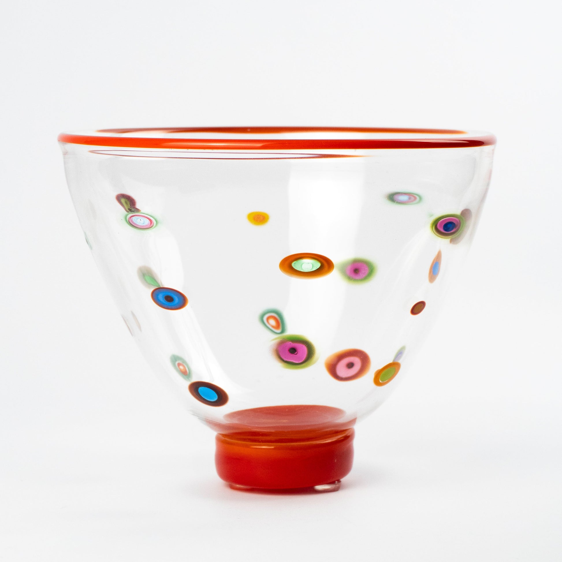 A clear glass bowl with a vibrant red rim and base, decorated with colorful hand-blown glass dots known as Murrine in various patterns, set against a plain white background.