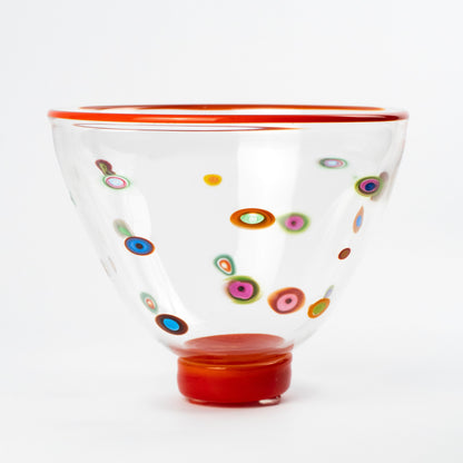 A clear glass bowl with a vibrant red rim and base, decorated with colorful hand-blown glass dots known as Murrine in various patterns, set against a plain white background.