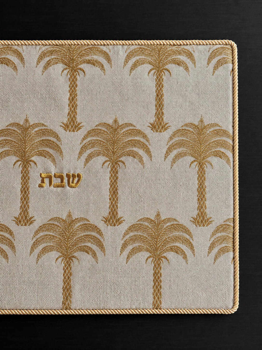 A rectangular challah cover with a cream linen fabric, gold palm tree pattern, and gold Hebrew lettering in the center reading shabbat, photographed from above on a dark wood surface.