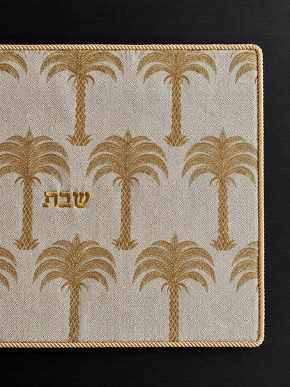 A rectangular challah cover with a cream linen fabric, gold palm tree pattern, and gold Hebrew lettering in the center reading shabbat, photographed from above on a dark wood surface.