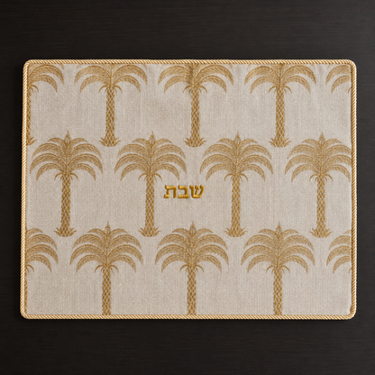 A rectangular challah cover with a cream linen fabric, gold palm tree pattern, and gold Hebrew lettering in the center reading shabbat, photographed from above on a dark wood surface.