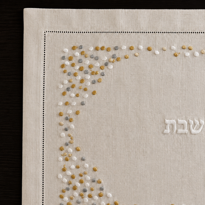 A cream linen Shabbat challah cover with a decorative border of gold and white embroidered dots and the Hebrew word 'Shabbat' in the center, displayed flat on a dark wood background.