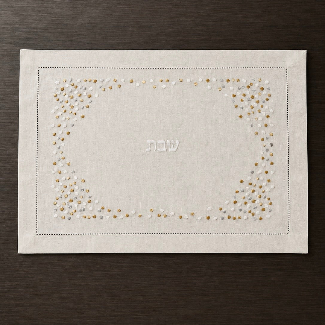 A cream linen Shabbat challah cover with a decorative border of gold and white embroidered dots and the Hebrew word 'Shabbat' in the center, displayed flat on a dark wood background.
