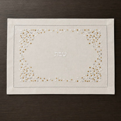 A cream linen Shabbat challah cover with a decorative border of gold and white embroidered dots and the Hebrew word 'Shabbat' in the center, displayed flat on a dark wood background.