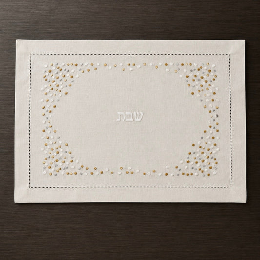A cream linen Shabbat challah cover with a decorative border of gold and white embroidered dots and the Hebrew word 'Shabbat' in the center, displayed flat on a dark wood background.