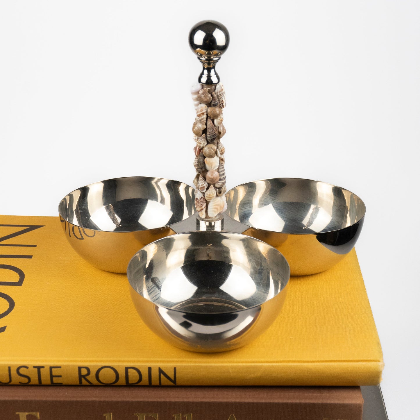 A polished silver-toned metal three-bowl serving stand with a decorative shell-wrapped handle and spherical finial, displayed on stacked books against a light gray background.