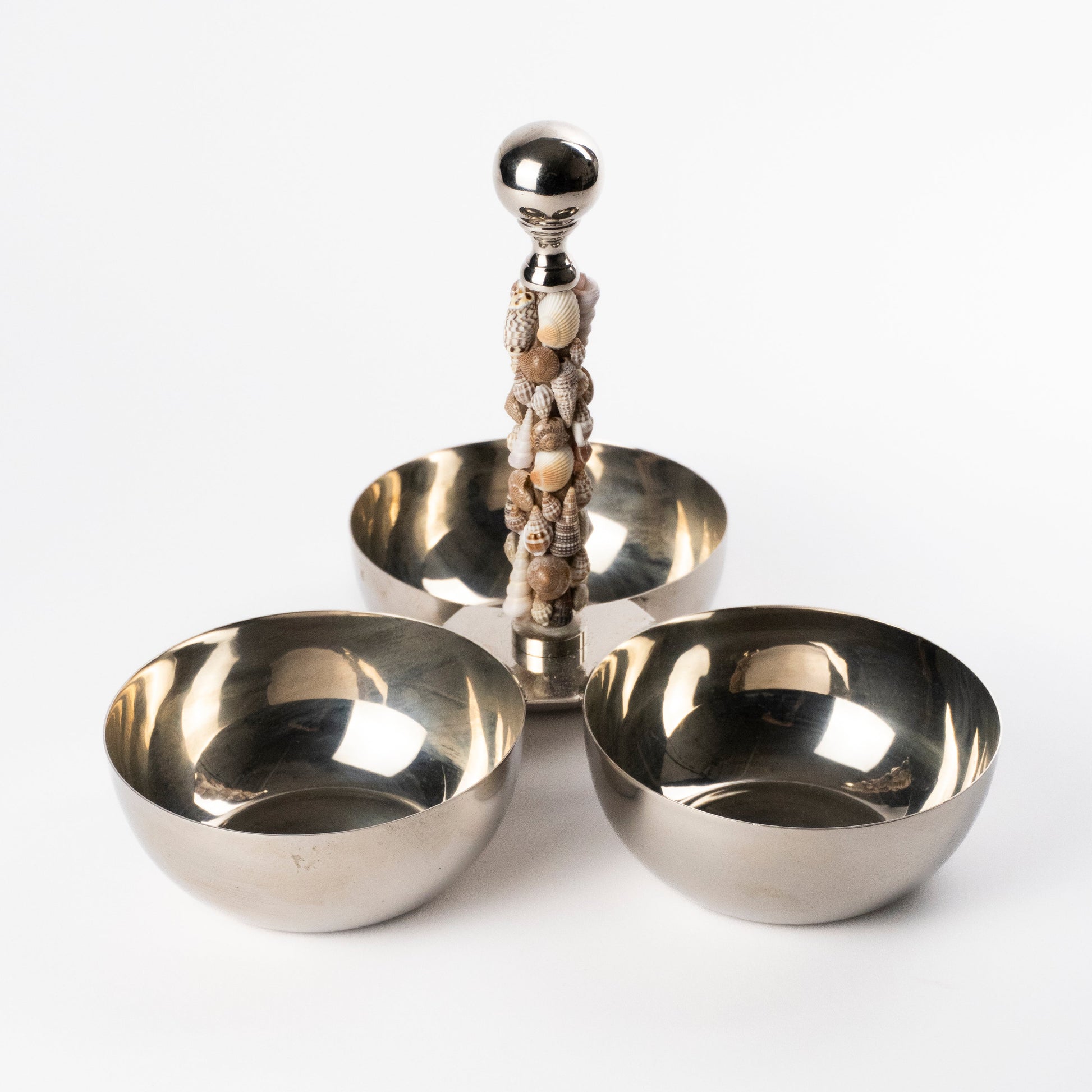 A polished silver-toned metal three-bowl serving stand with a central pillar designed with seashells, photographed on a clean white studio background.