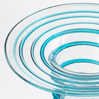 A close-up of a clear glass vase with concentric spiral ridges and a flared top, showing bright turquoise blue tint through the transparent glass against a white background.