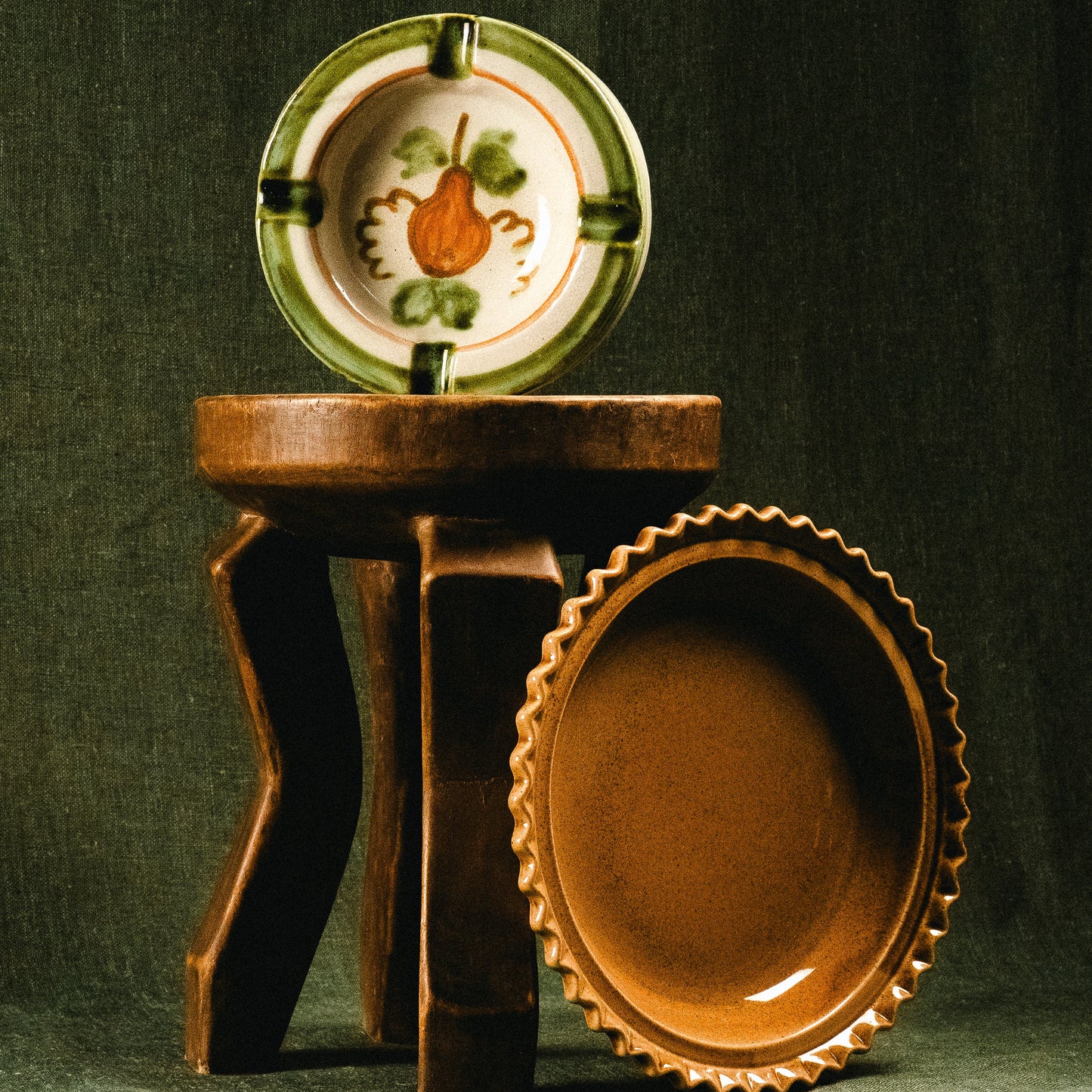 A rustic ceramic bowl with a scalloped rim and warm brown interior, displayed alongside a matching decorative plate on a dark wooden stand against a dark green backdrop.