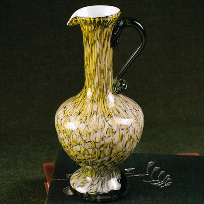 A decorative glass pitcher with a yellow and white marbled pattern and a black handle, displayed on top of two dark books against a dark green background.