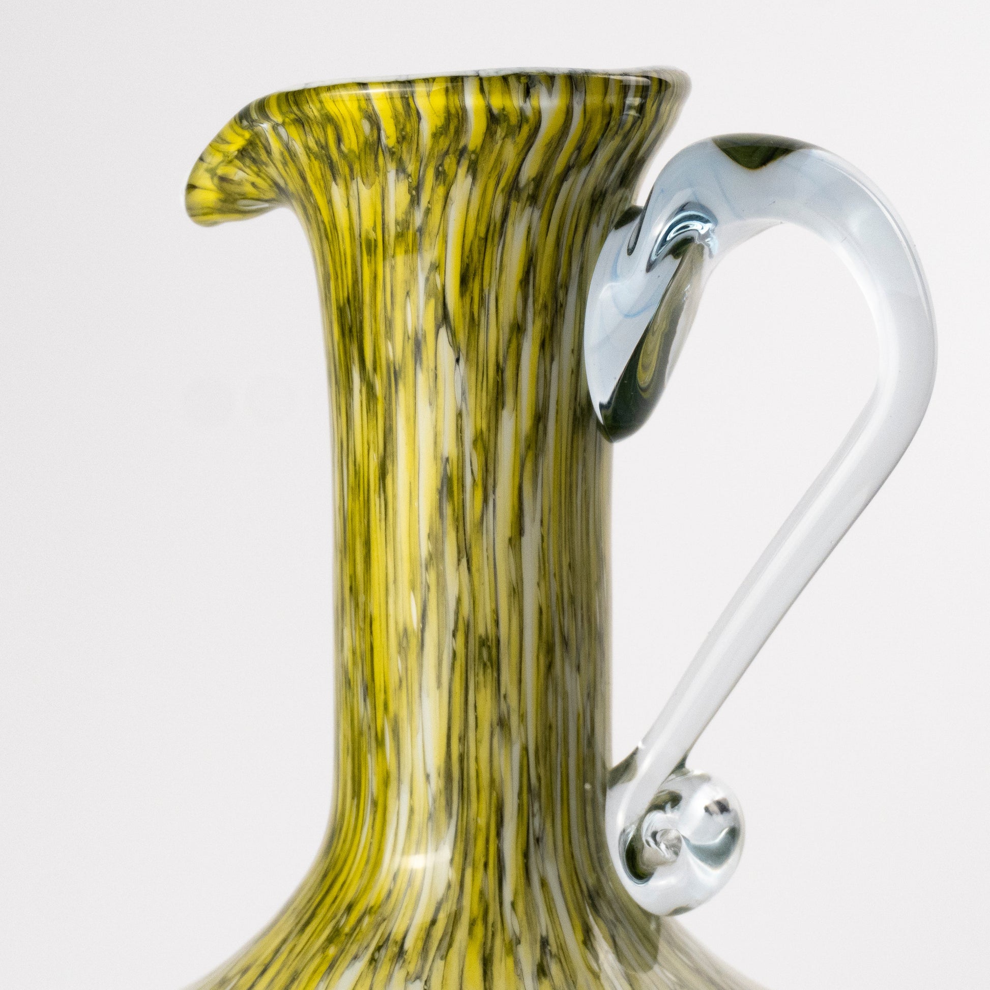 Close-up of a decorative glass pitcher with a flared neck, vertical marbled yellow and olive-green pattern, and a curved clear glass handle on a light gray background.