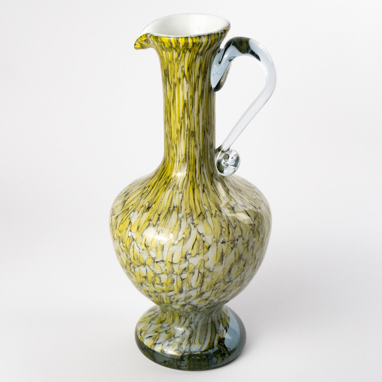 A decorative murano glass pitcher with a bulbous body, flared base, and curved handle, featuring a marbled pattern of olive green, yellow, and white swirls with a clear glass handle.