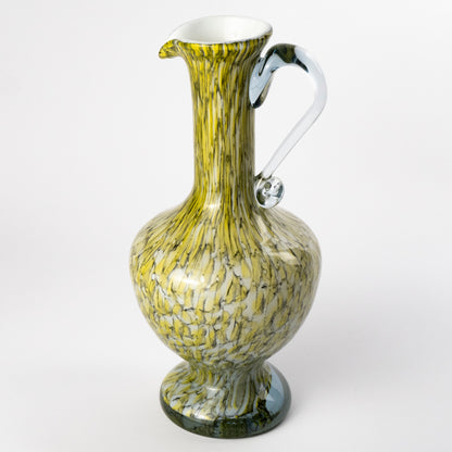 A decorative murano glass pitcher with a bulbous body, flared base, and curved handle, featuring a marbled pattern of olive green, yellow, and white swirls with a clear glass handle.