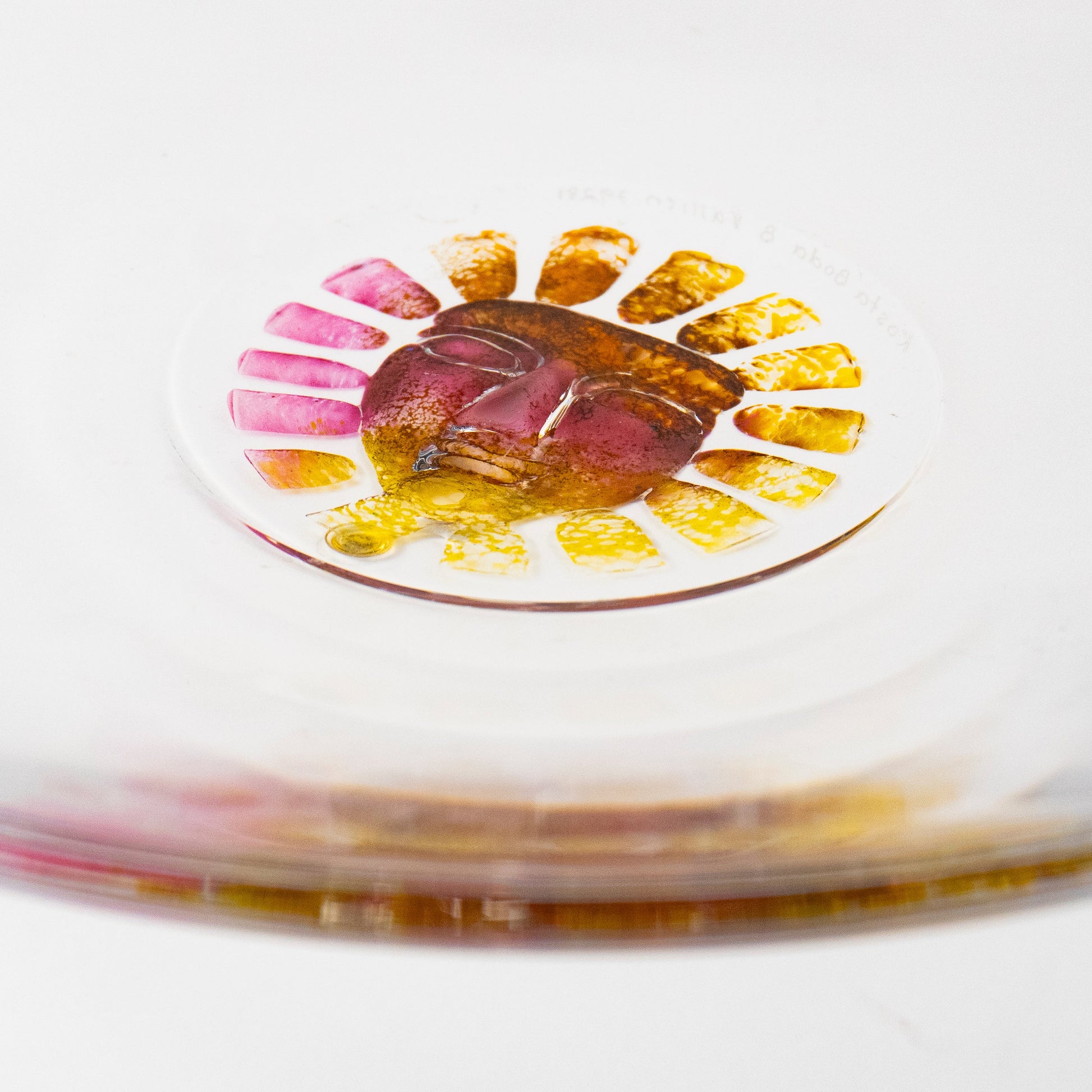 A close-up of a clear circular glass dish or plate resting on a brushed silver metal surface, with a small colorful floral sun inset containing pink, yellow, and orange details.