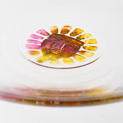 A close-up of a clear circular glass dish or plate resting on a brushed silver metal surface, with a small colorful floral sun inset containing pink, yellow, and orange details.