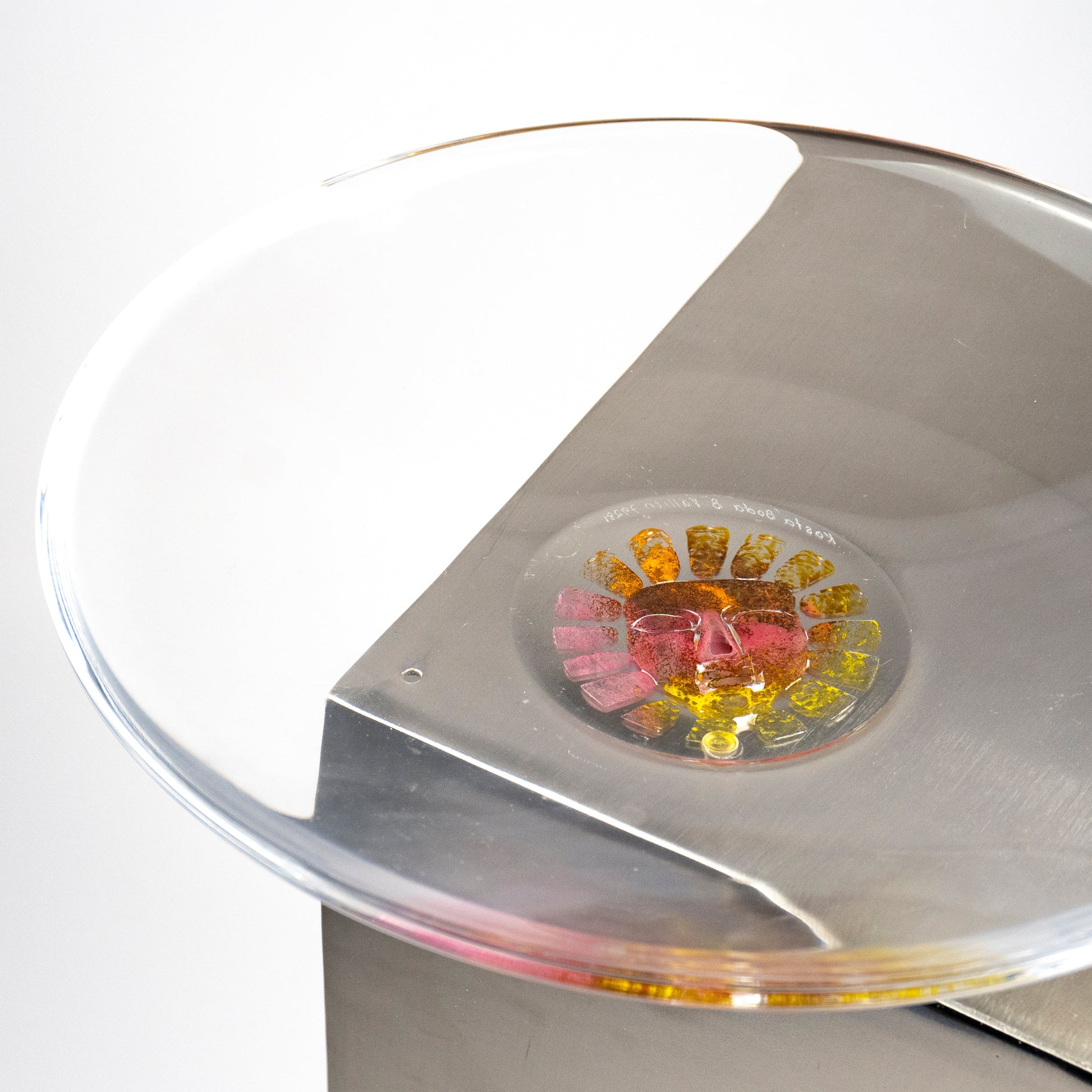 A side view of a clear circular glass dish or plate resting on a brushed silver metal surface, with a small colorful floral sun inset containing pink, yellow, and orange details.