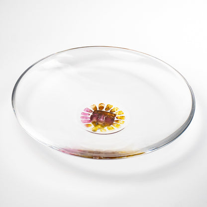A clear glass shallow dish with a small hand-painted floral sun motif in the center, featuring pink, yellow, and purple details, set against a clean white background.