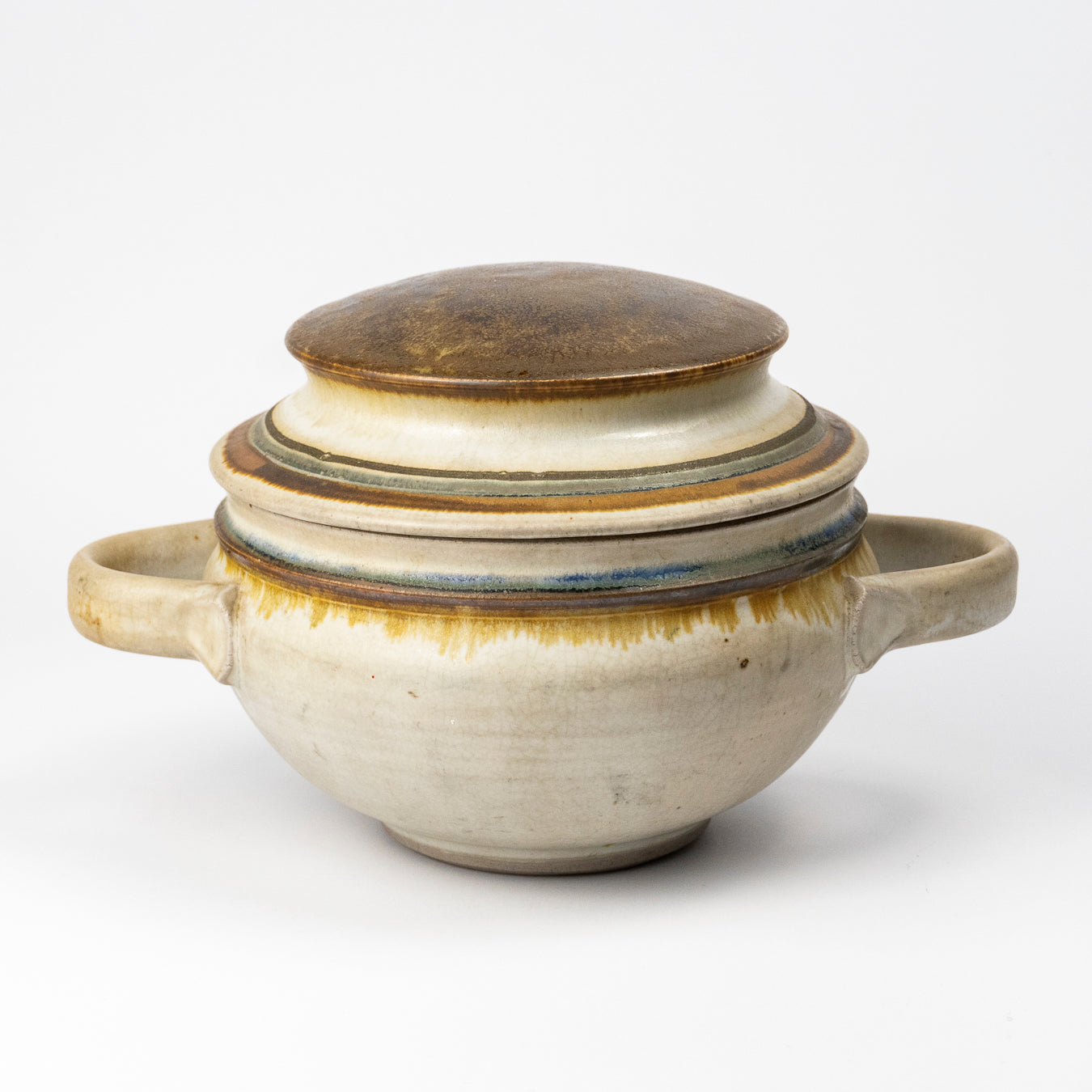A handmade ceramic lidded soup or stew pot with side handles, featuring a textured cream and beige glaze with a brown top lid and subtle blue and yellow-green glaze accents.