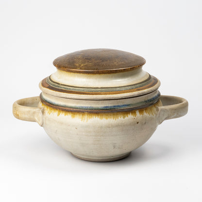 A handmade ceramic lidded soup or stew pot with side handles, featuring a textured cream and beige glaze with a brown top lid and subtle blue and yellow-green glaze accents.
