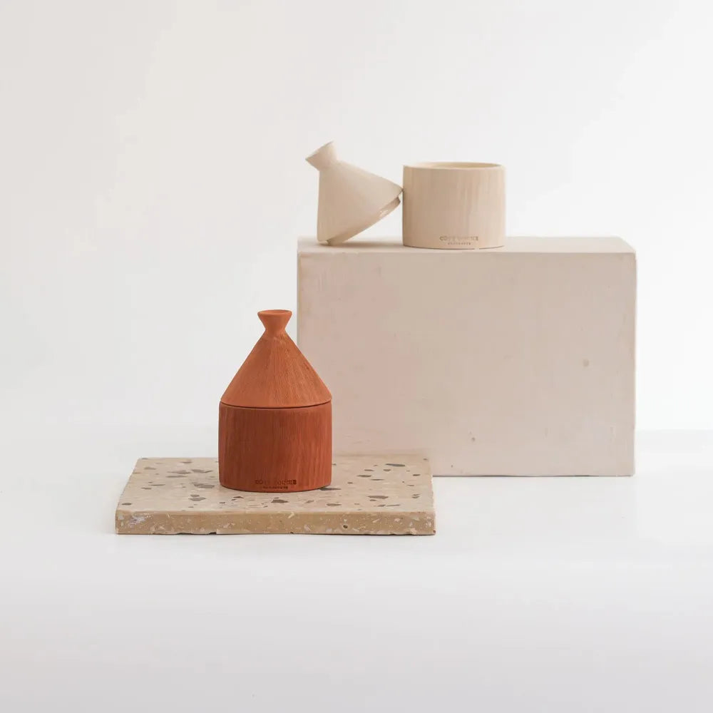 Minimalist terracotta and cream ceramic objects arranged on a white studio background, including a small cone-shaped vessel on a speckled stone slab and a larger cream block with two cream ceramic pieces on top.
