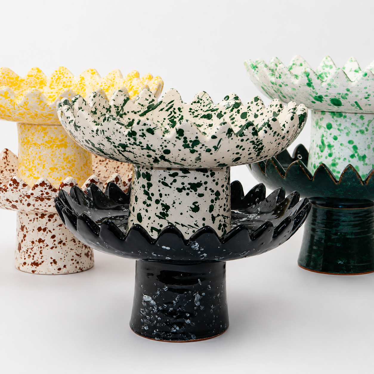 Floree ceramic pedestal bowls in five colorways — yellow, brown, green, black, and deep green speckle — with sawtooth zigzag rims and footed bases
